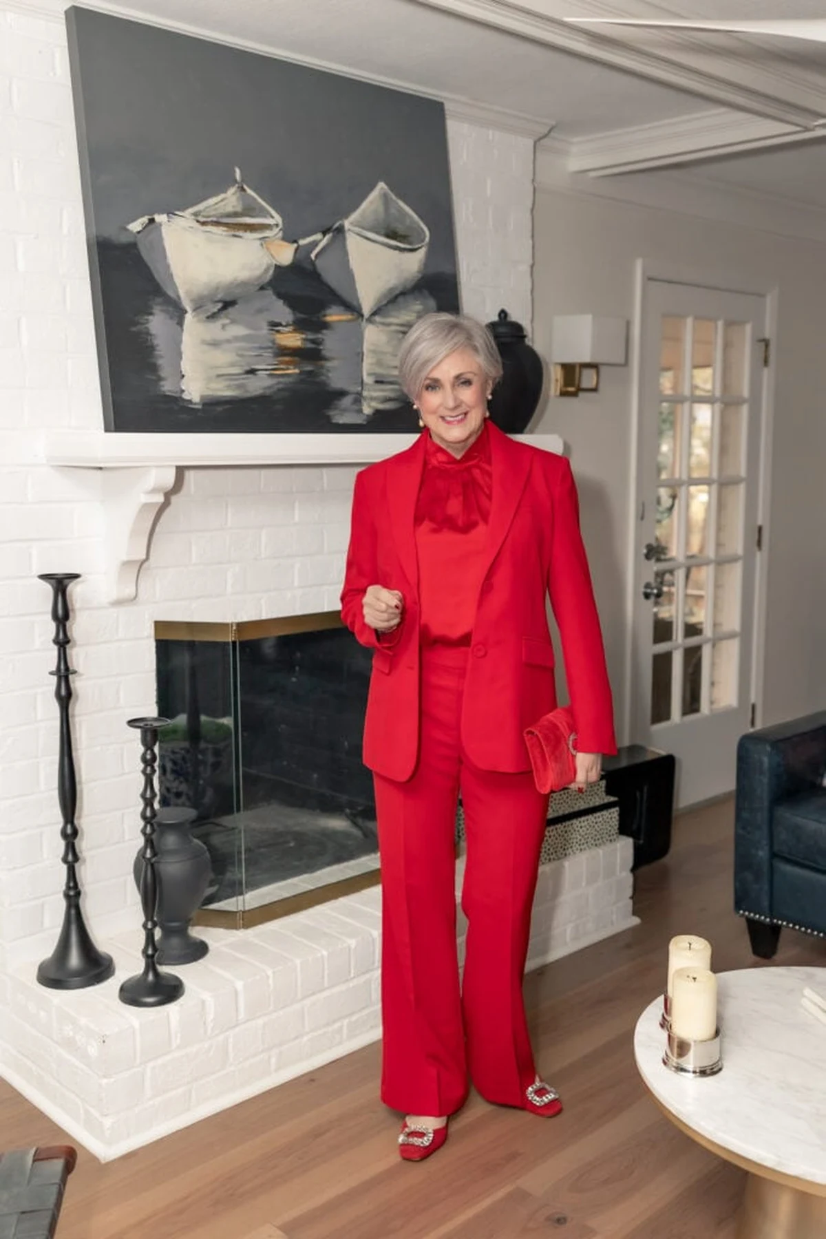 A woman over 50 wears an all-red power suit with an open blazer beside a white-brick fireplace decorated for Christmas.