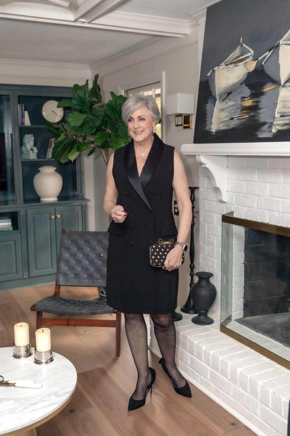 A woman over 50 wears a black tuxedo dress with satin lapels and patterned tights in front of a Christmas fireplace.