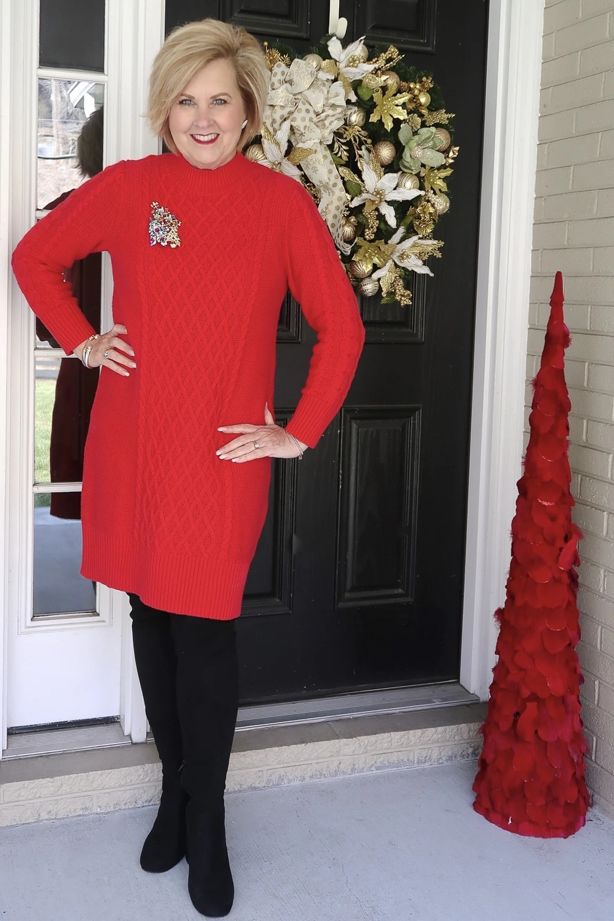 A woman over 50 wears a red cable-knit sweater dress with a jeweled tree brooch and over-knee boots at a Christmas-decorated door.