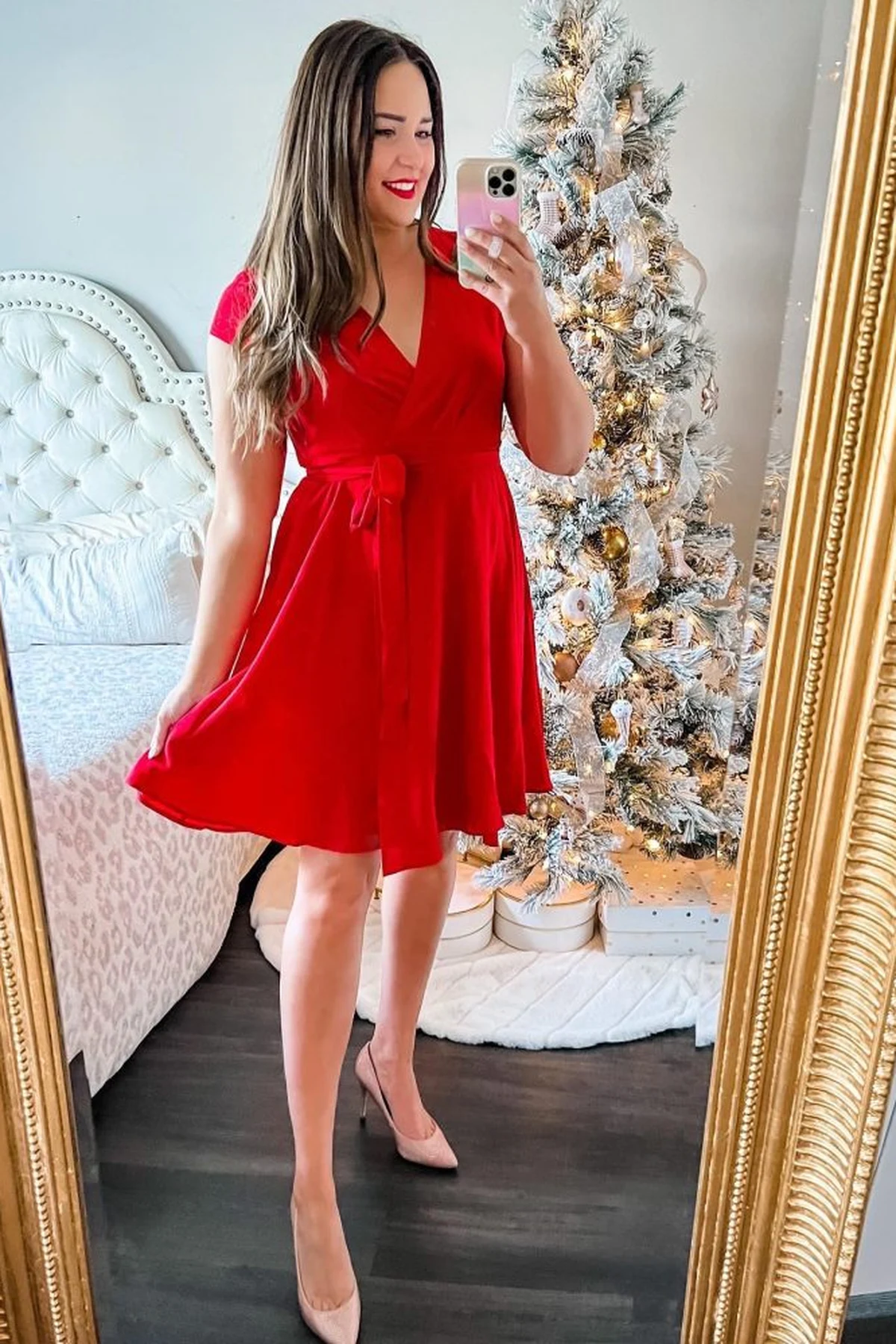 A woman over 50 takes a mirror selfie in a red V-neck wrap dress in front of a lit flocked Christmas tree.