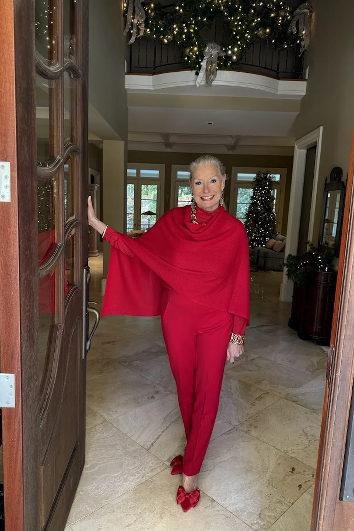 A woman over 50 wears a red draped poncho with slim trousers in a foyer with a Christmas tree and garlands.