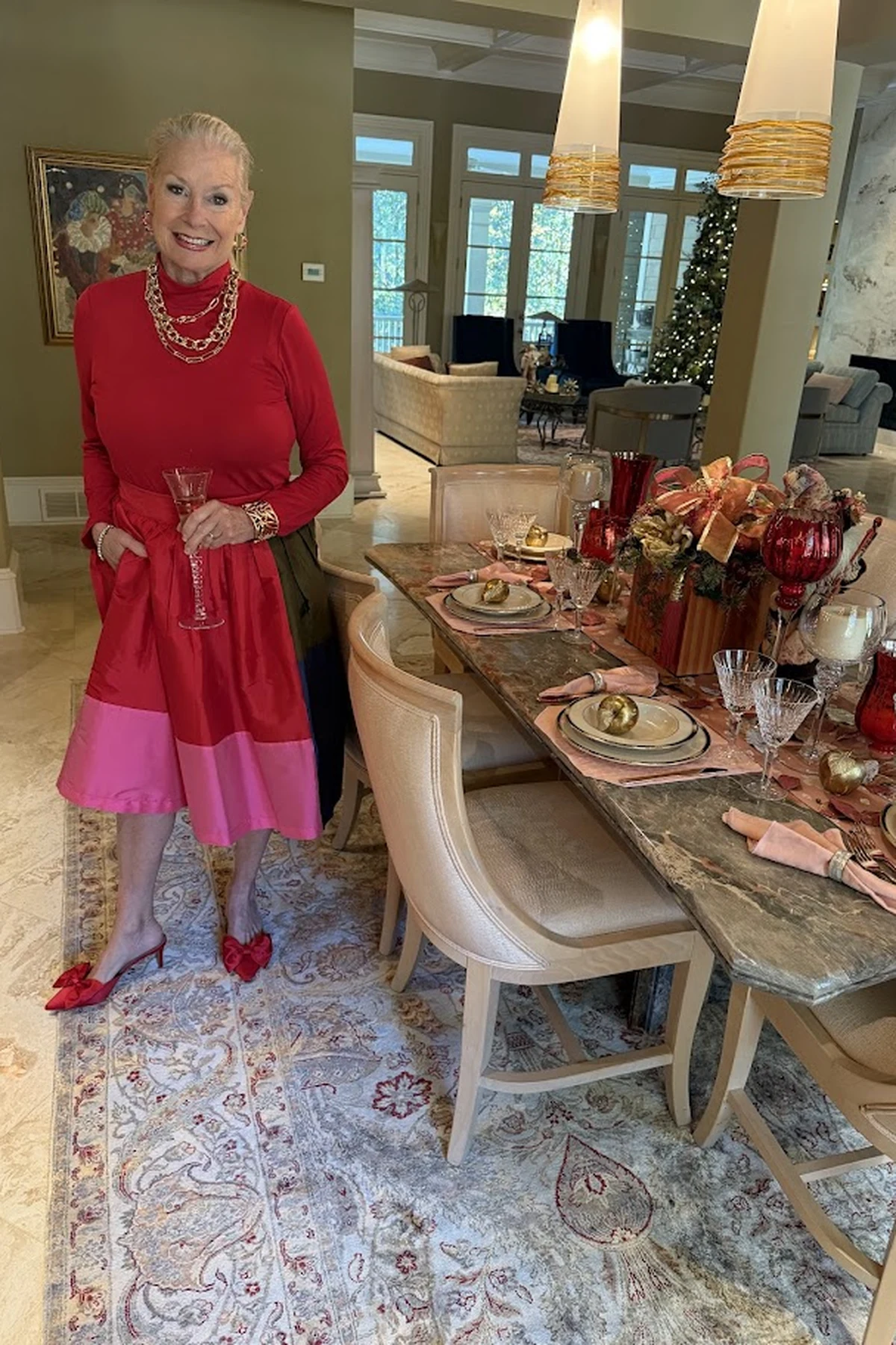 A woman over 50 wears a red turtleneck and colorblock midi skirt holding champagne at a Christmas dining table.