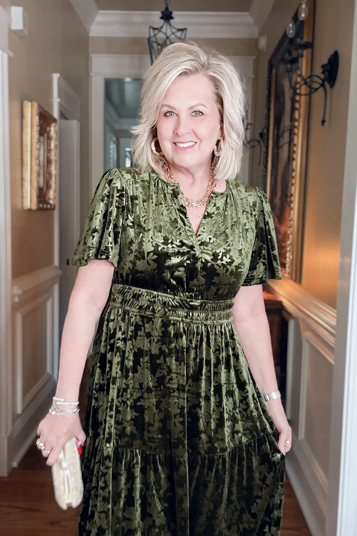 A woman over 50 wears an olive-green floral velvet empire-waist dress with gold jewelry.