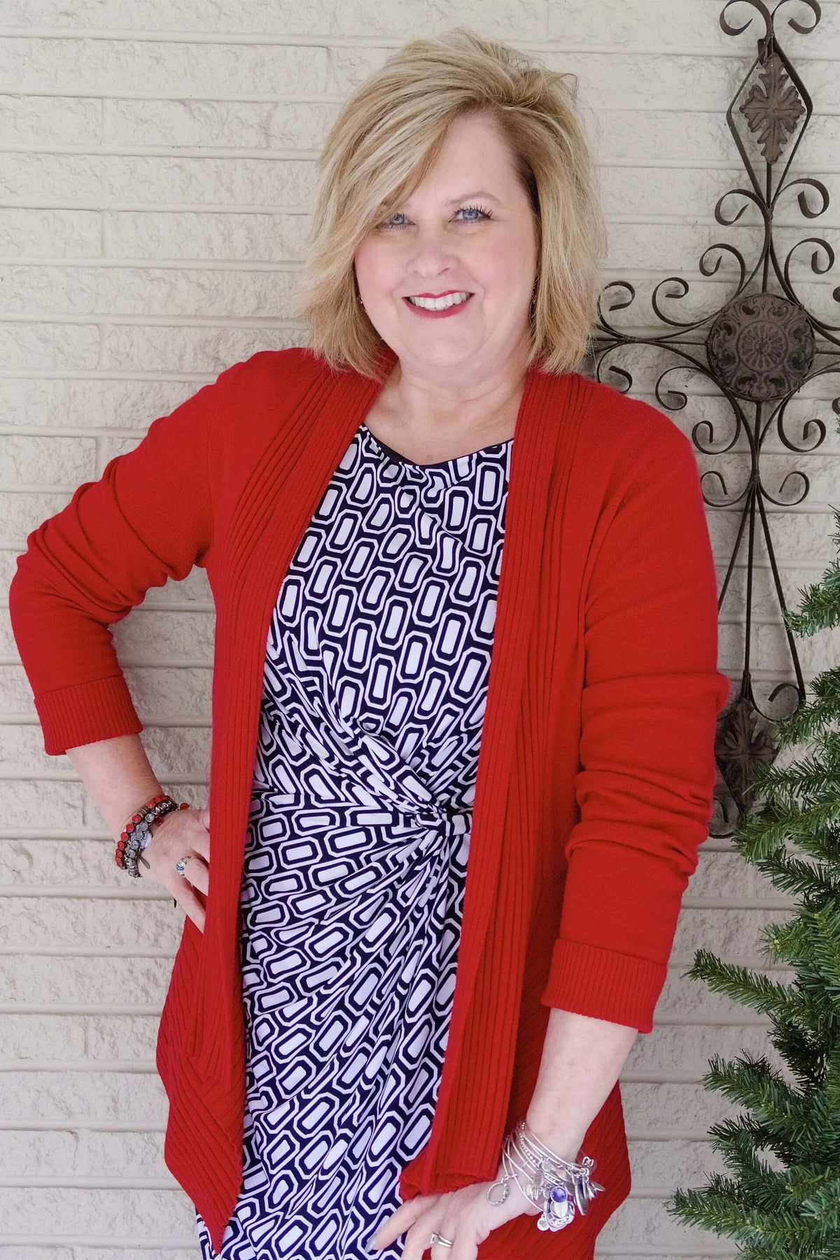 A woman over 50 wears a red open cardigan over a geometric wrap dress beside a Christmas tree.