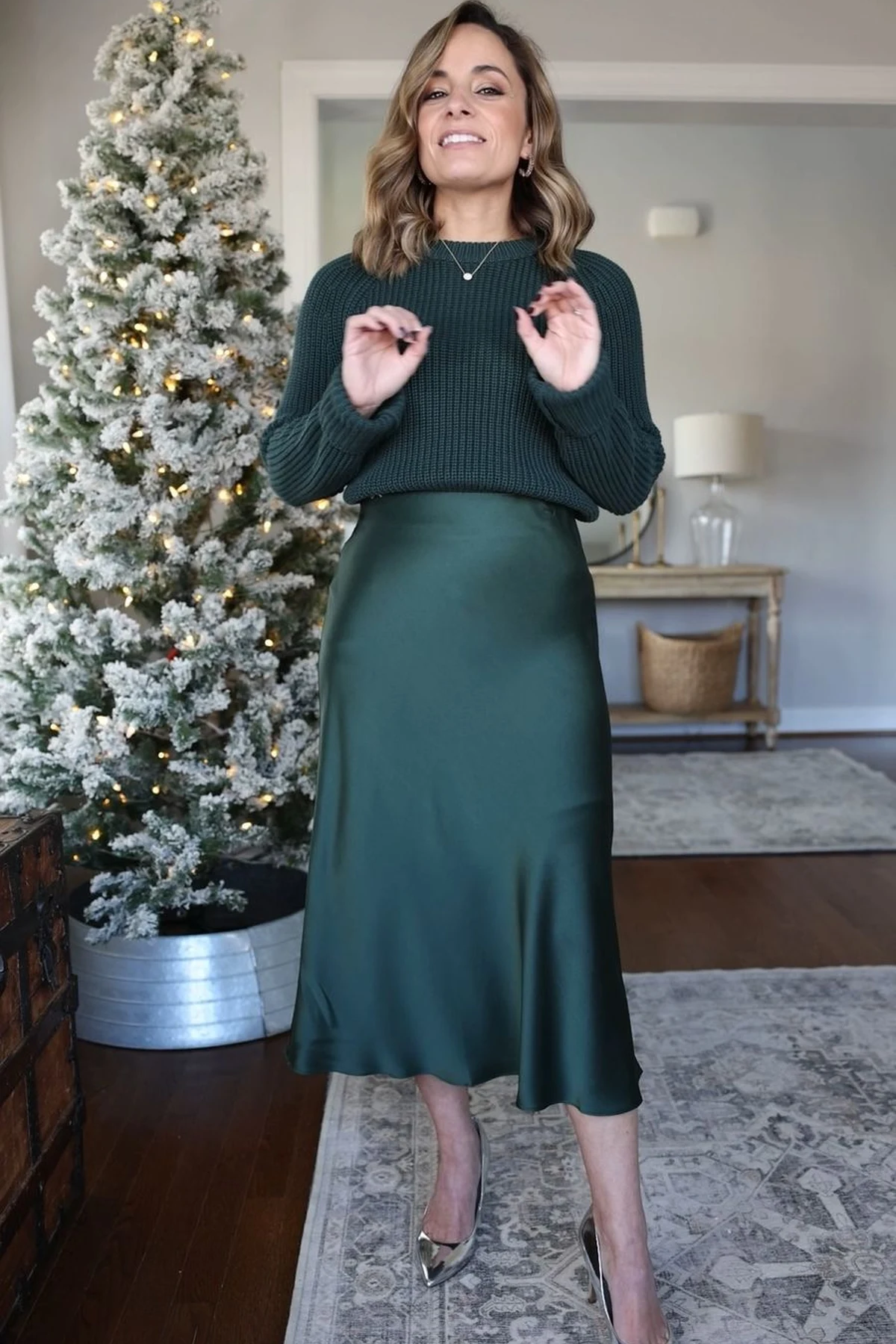 A woman wears a green chunky-knit sweater with an emerald satin midi skirt beside a flocked Christmas tree.