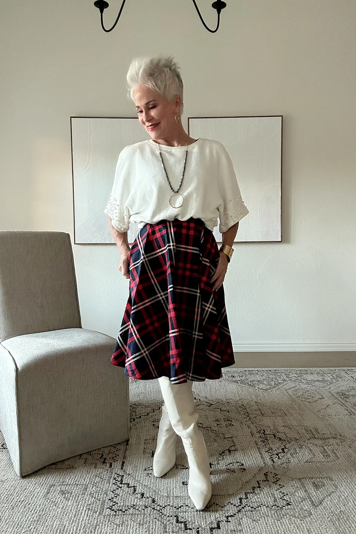 A woman over 50 wears a white sweater with a plaid A-line midi skirt and white knee boots.