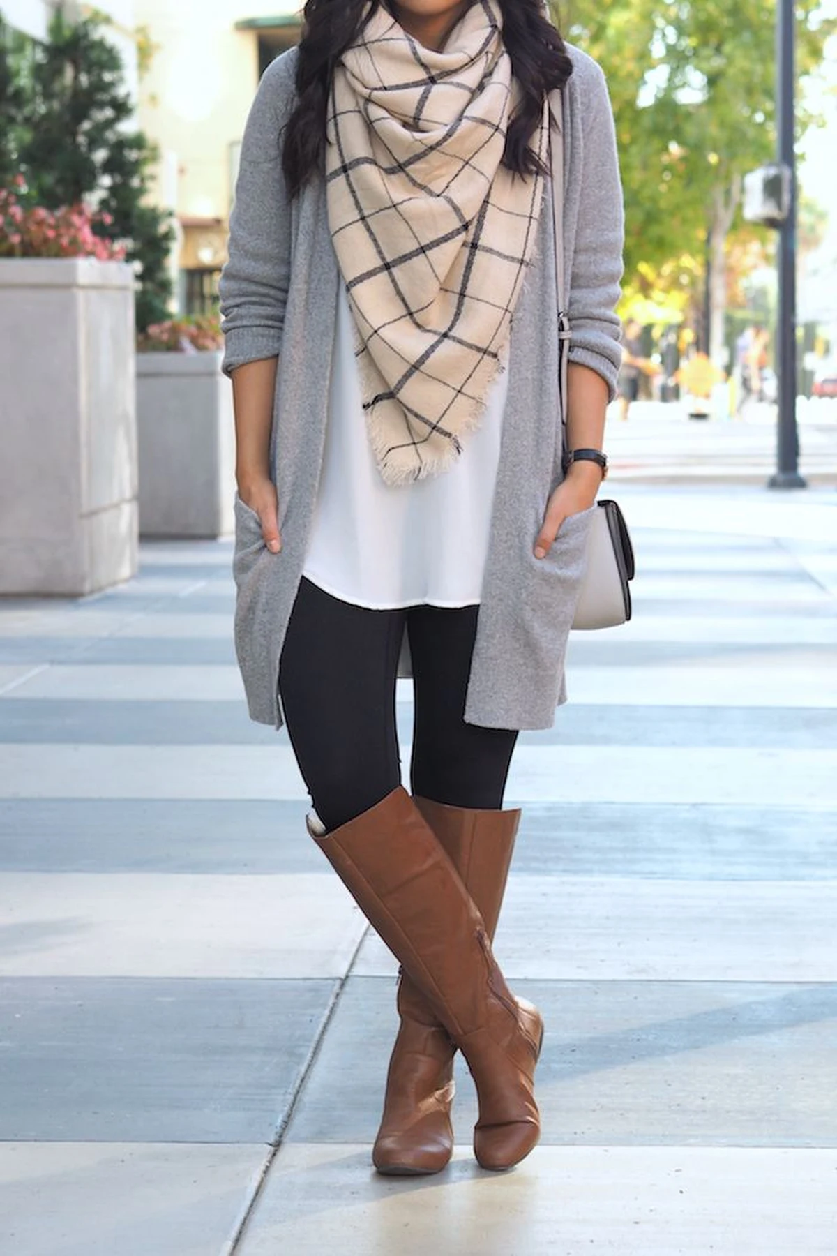 A woman wears a long grey cardigan with a plaid blanket scarf, black leggings and knee-high boots.