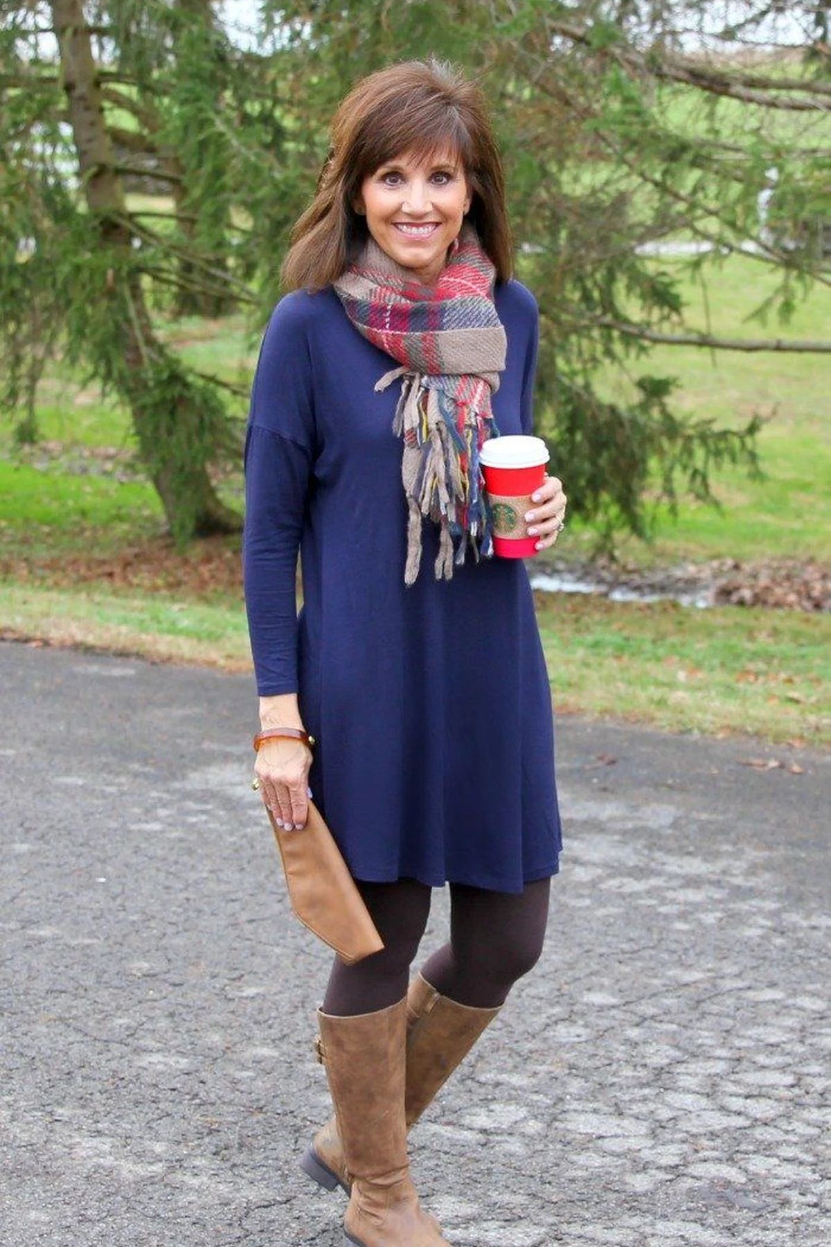 A woman wears a navy tunic with a red plaid scarf and tan knee boots among evergreen trees.