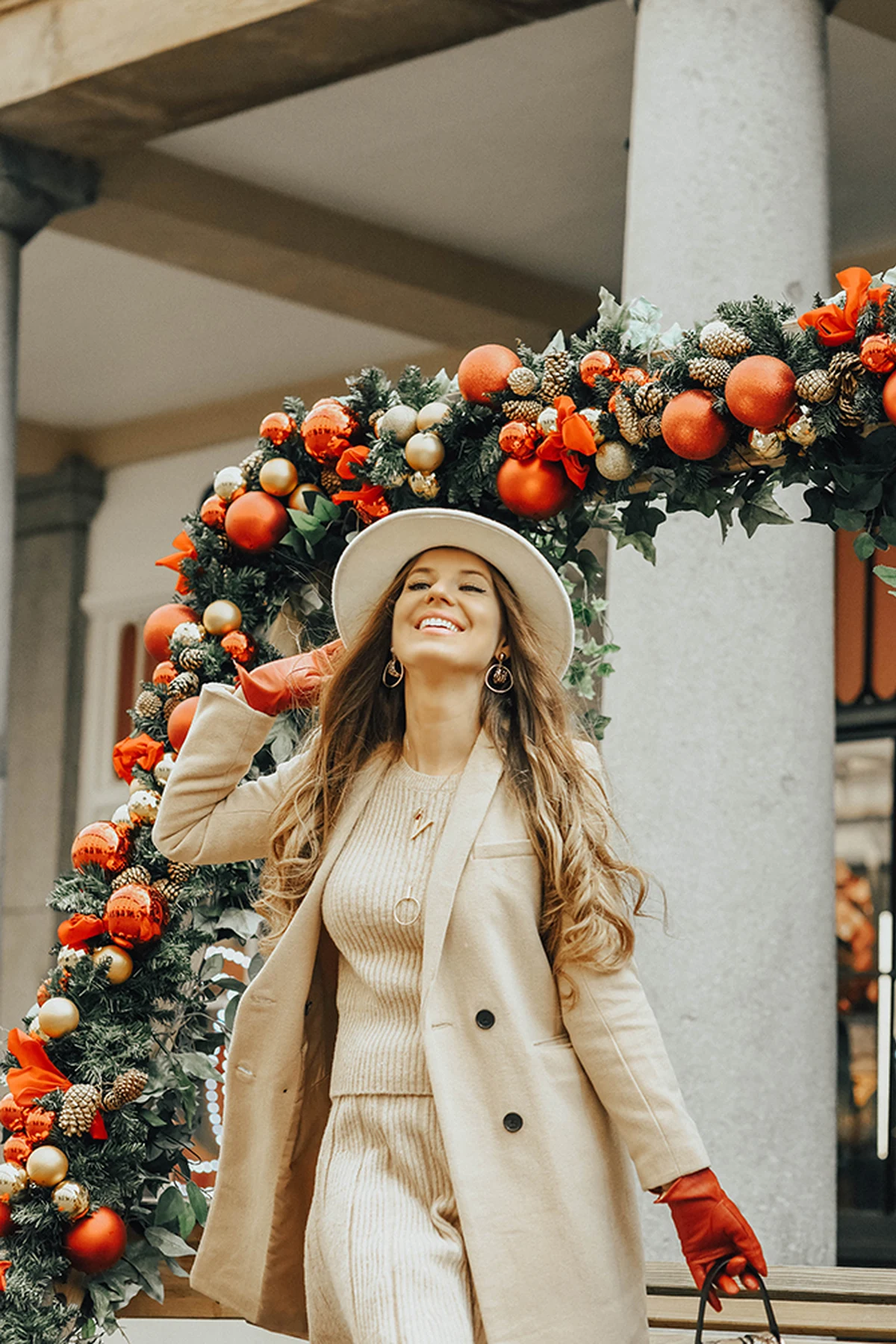 A woman over 50 wears a cream cable-knit dress with a camel coat and red gloves under a Christmas wreath arch.