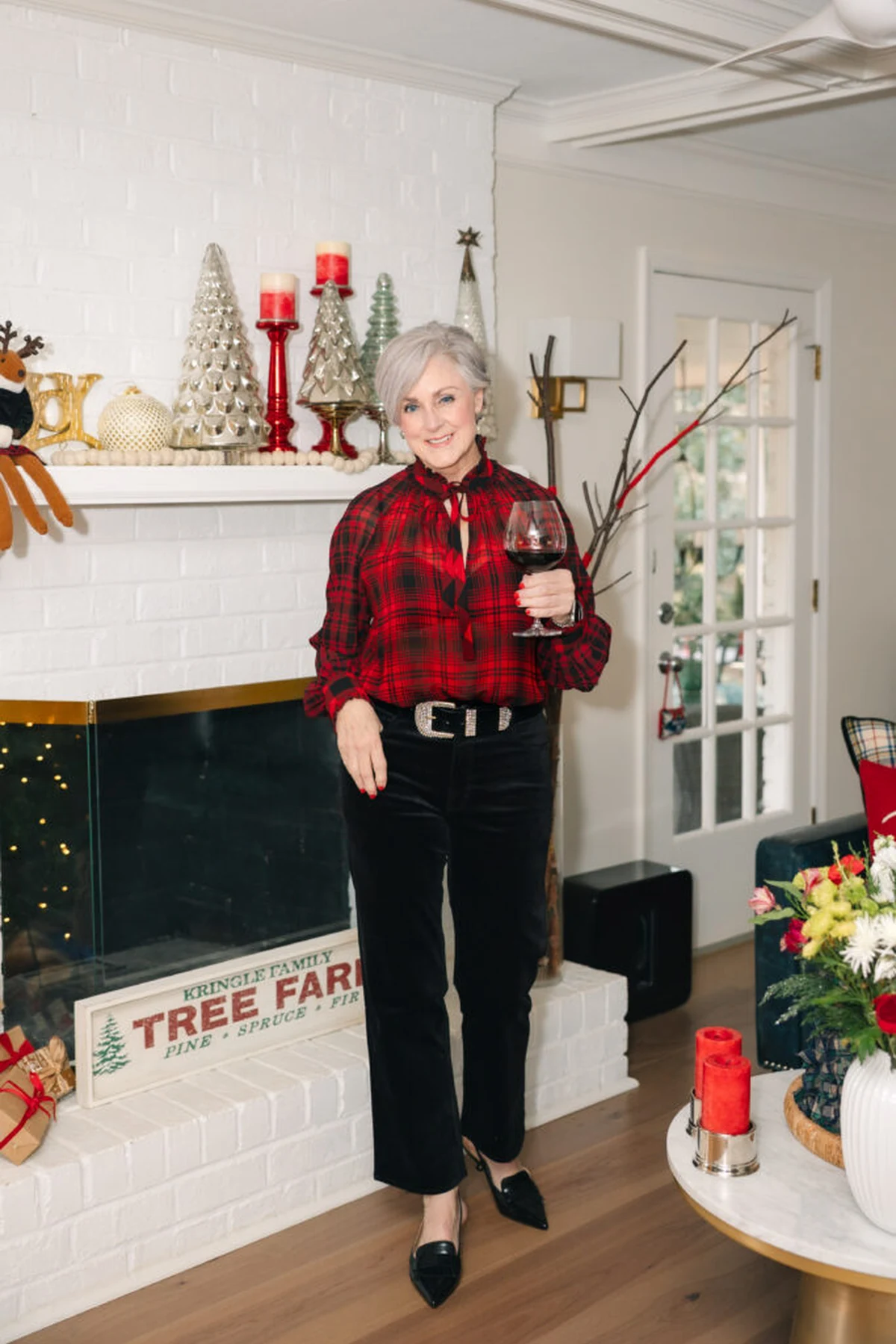 A woman over 50 wears a plaid tie-neck blouse with black velvet trousers by a Christmas fireplace.