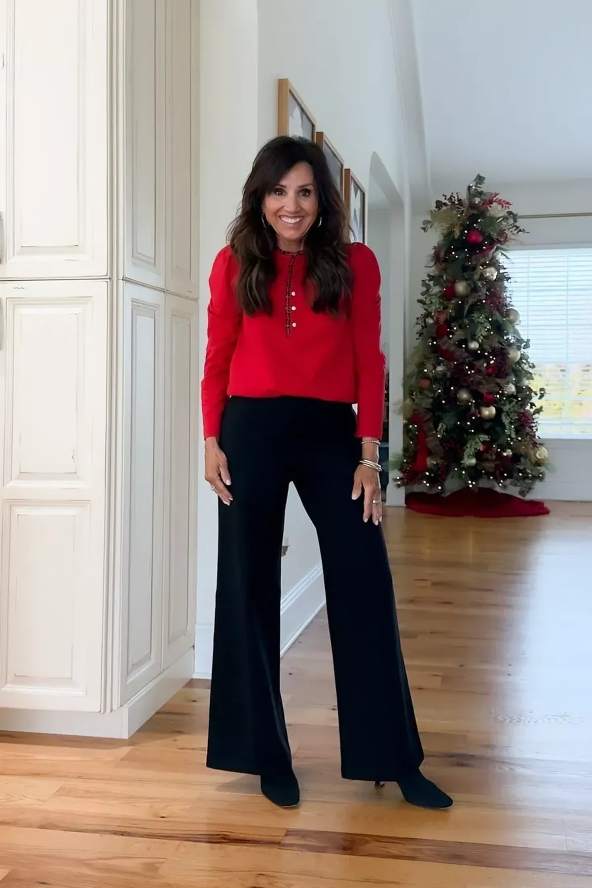 A woman wears a red henley blouse with black wide-leg trousers in front of a Christmas tree.