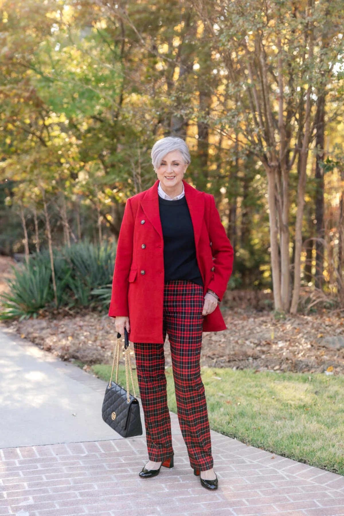 A woman over 50 wears a red coat worn open over a black top with tartan trousers.