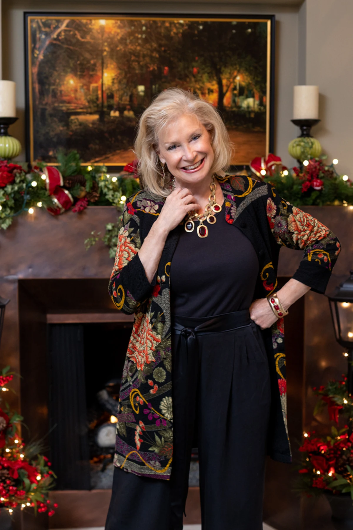 A woman over 50 wears a floral open-front jacket over black wide-leg trousers by a Christmas fireplace.
