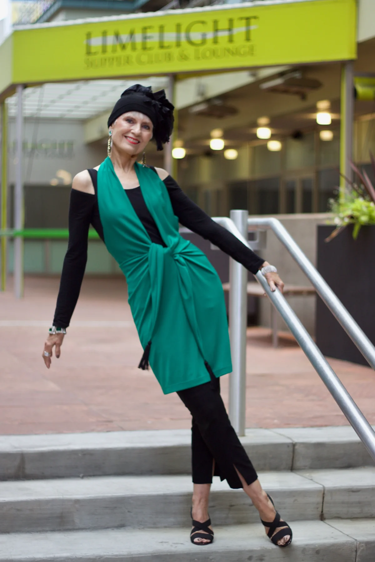 A woman over 50 wears an emerald twist-front tunic with black slim trousers.