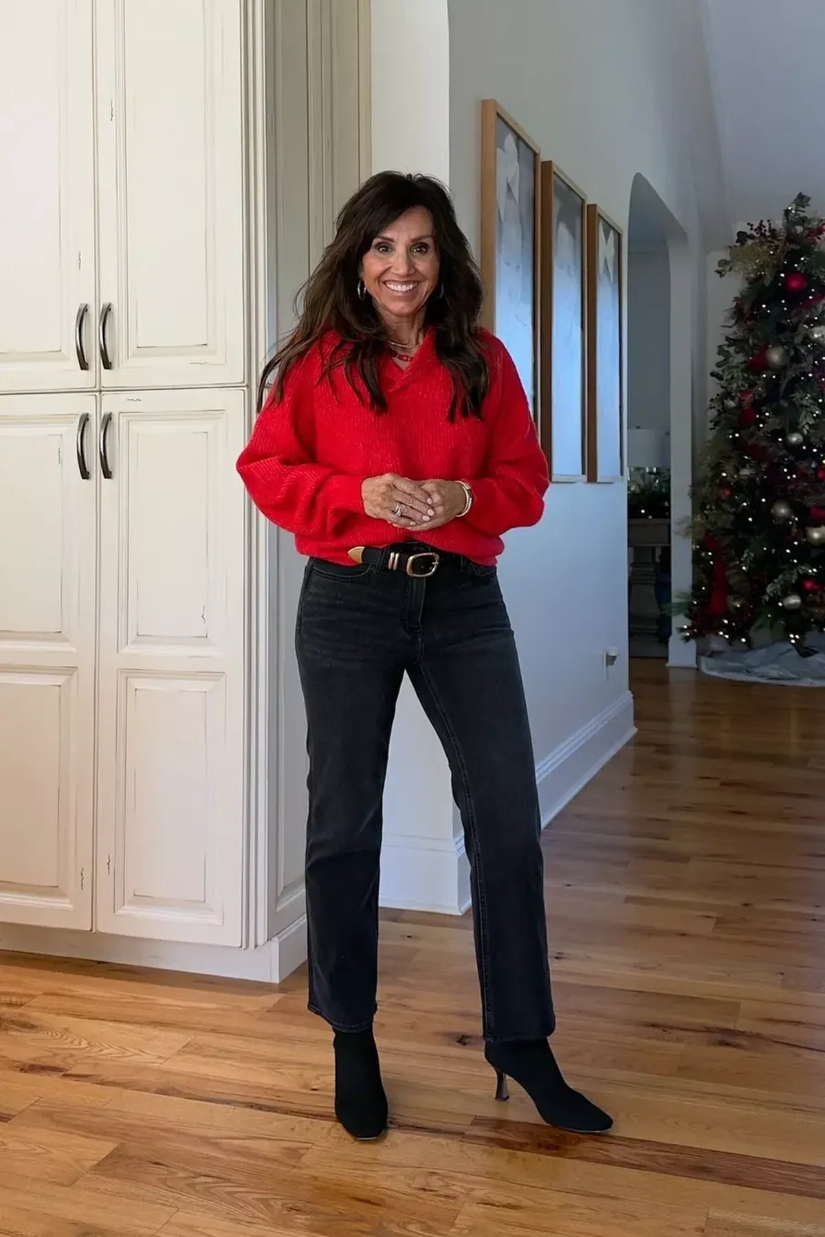 A woman wears a red V-neck sweater with black jeans and heeled booties in front of a Christmas tree.