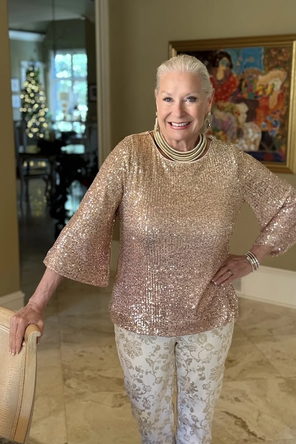 A woman over 50 wears a gold sequin top with brocade trousers in front of a lit Christmas tree.