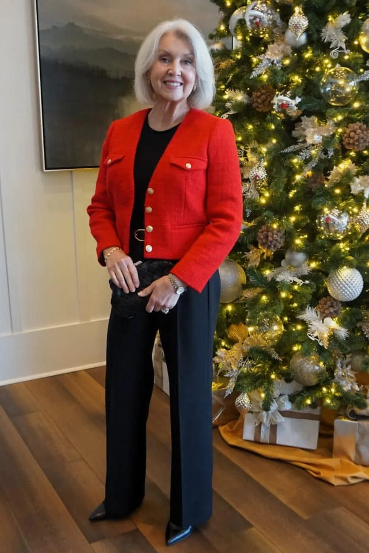 A woman over 60 wears a red tweed jacket with gold buttons over black beside a decorated Christmas tree.