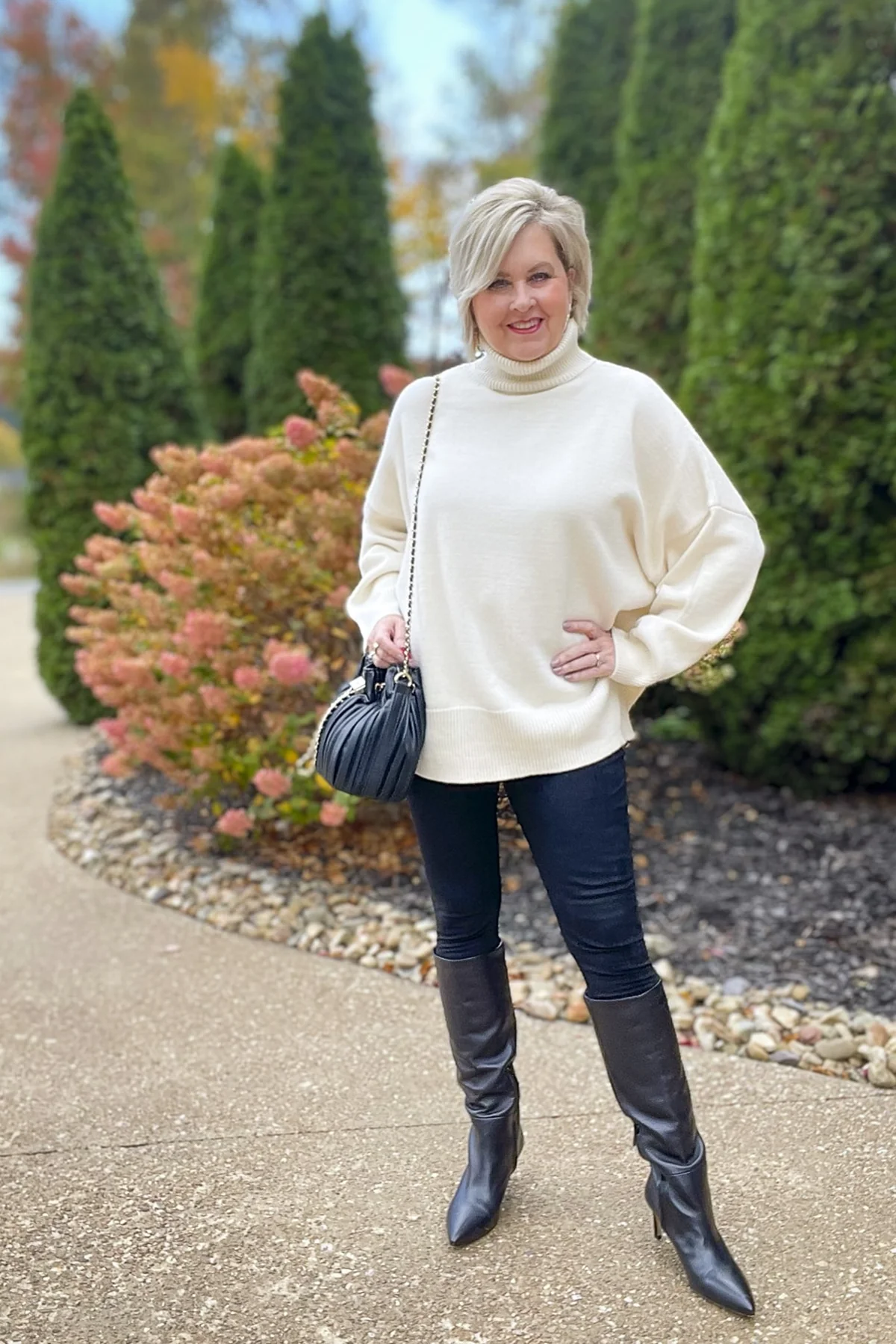 A woman over 50 wears an oversized cream turtleneck with black leggings and knee boots among evergreens.