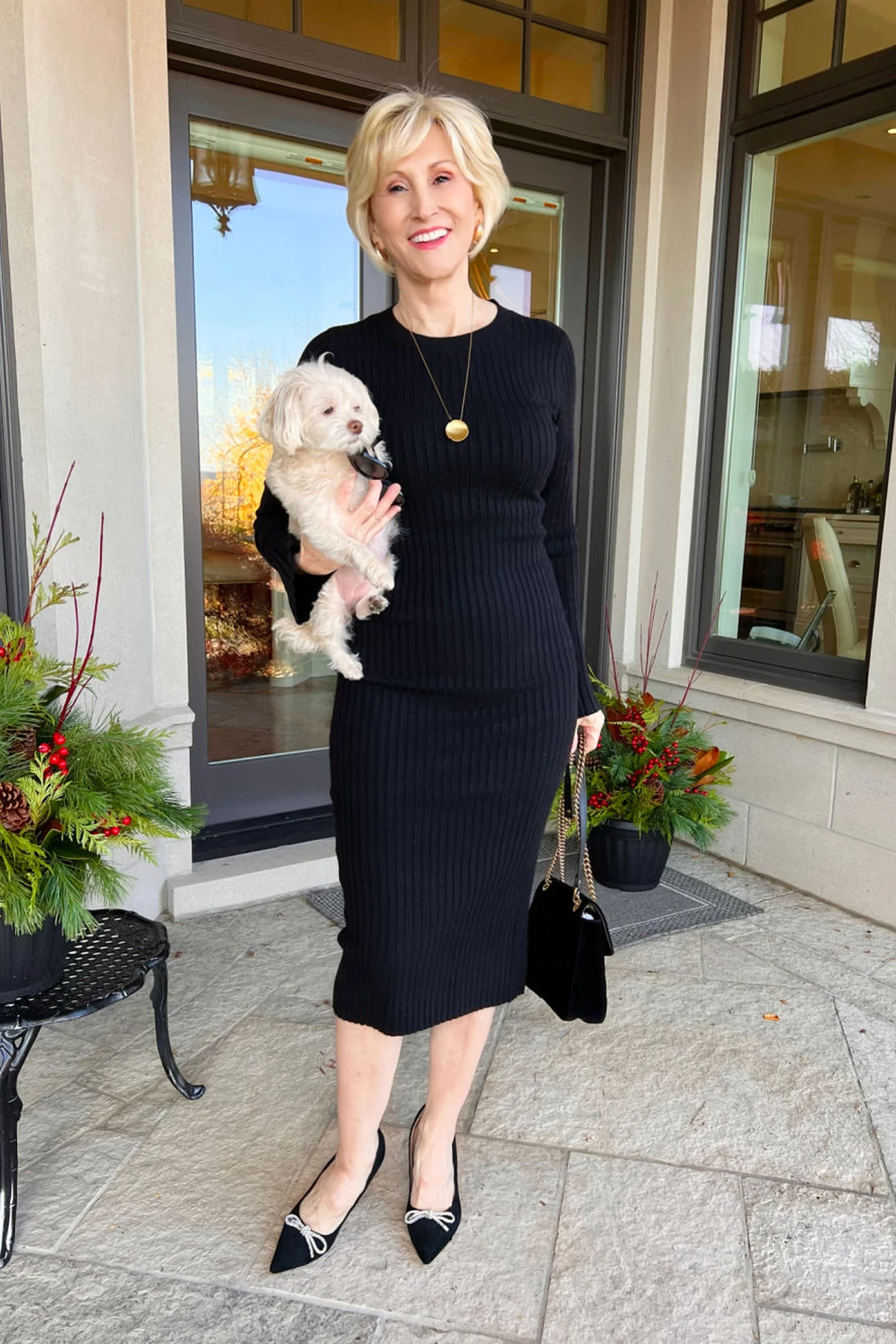 Woman in black ribbed-knit midi dress with gold pendant on a porch with holiday greenery