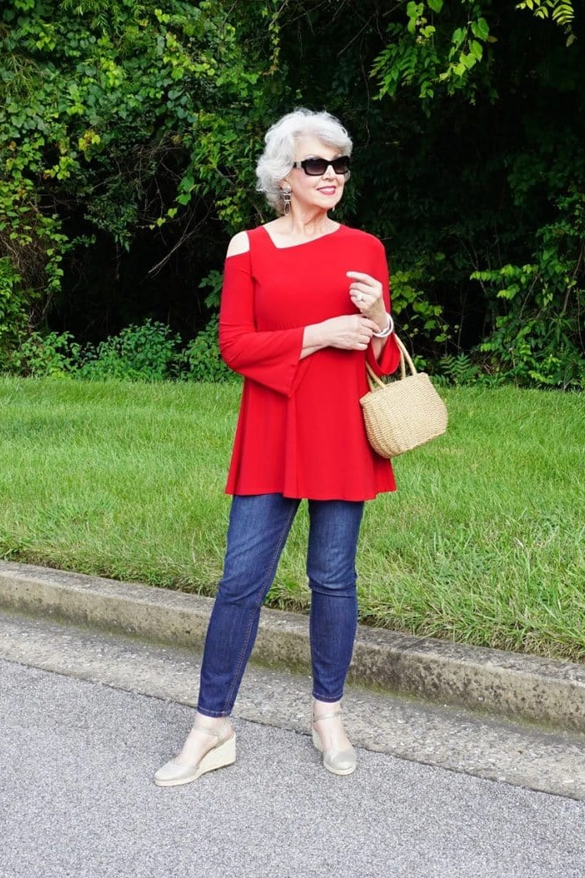 Woman in red peplum tunic and dark jeans on a roadside with green foliage