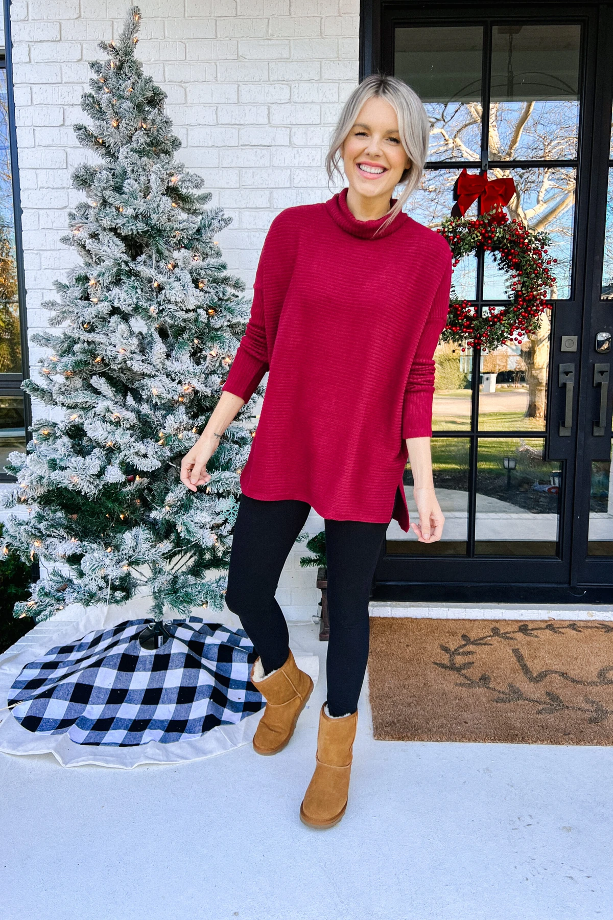 Woman in red tunic sweater and black leggings on a porch with a lit Christmas tree