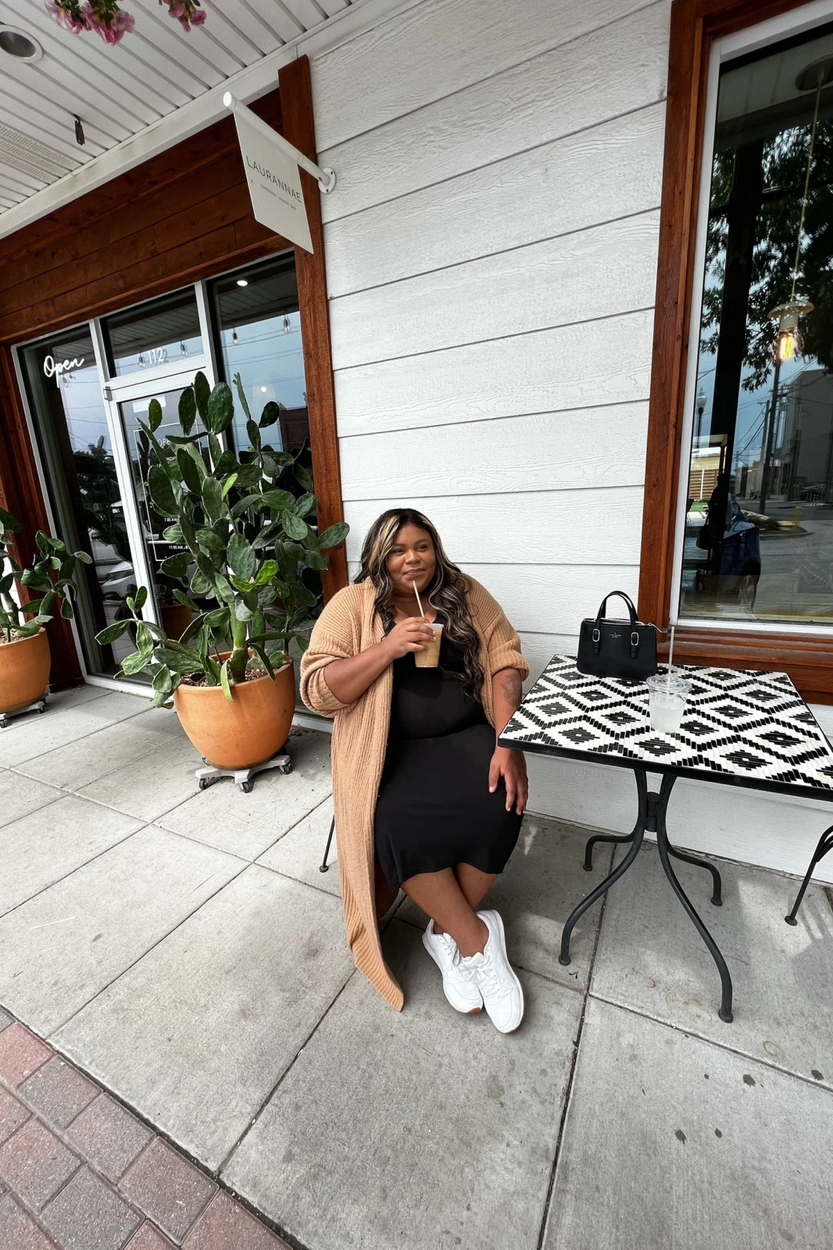 Woman in camel duster cardigan and black midi dress seated outside a cafe