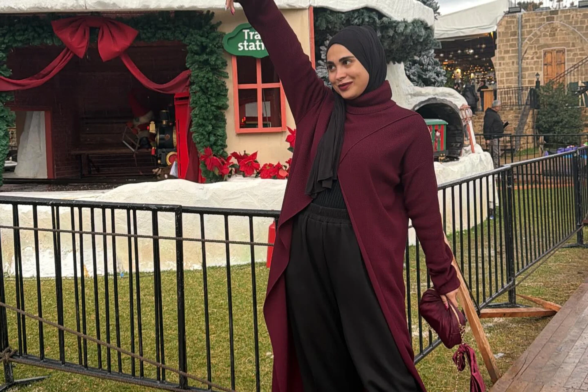 Hijabi woman in a maroon cardigan and black hijab at a Christmas-decorated train station