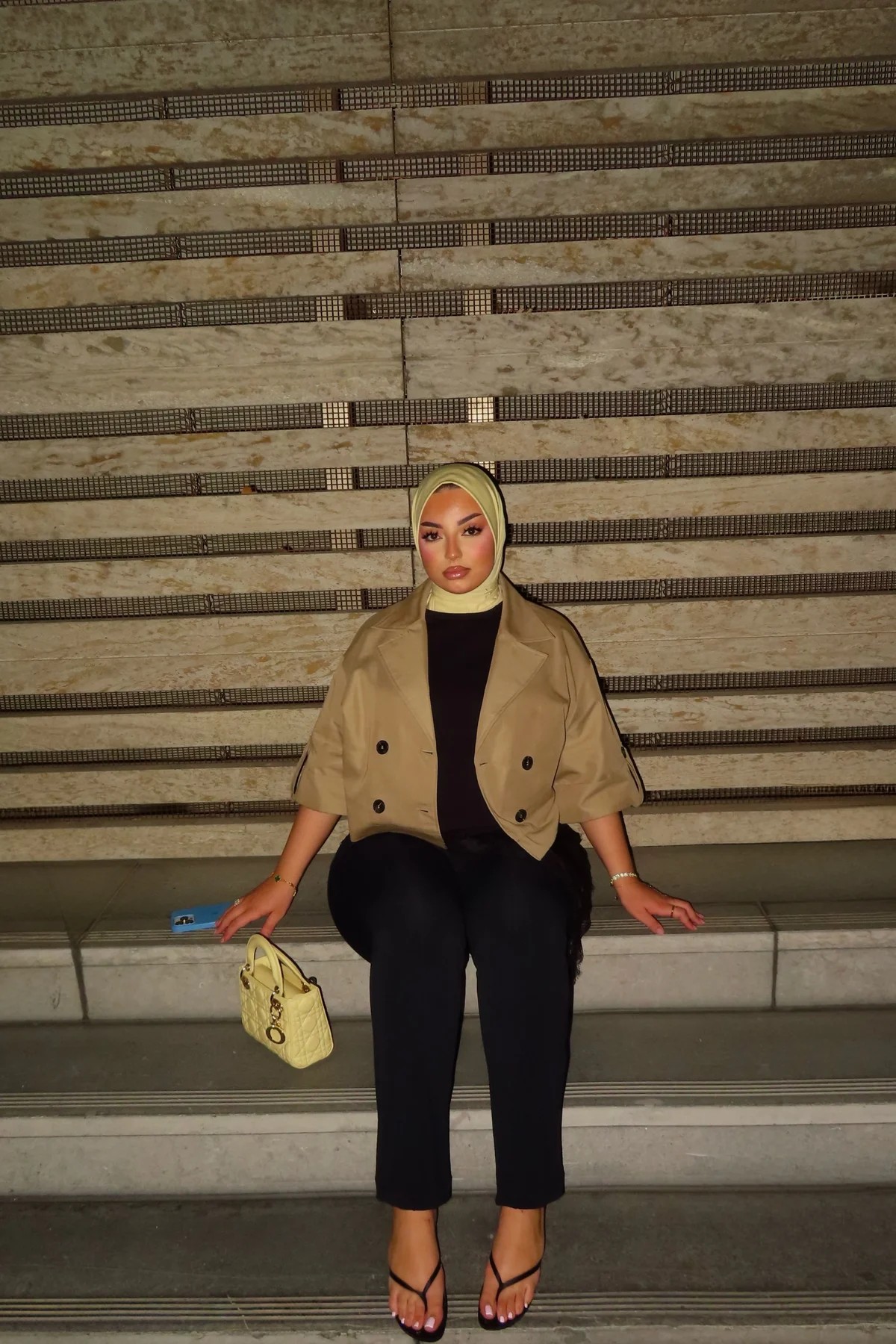 Hijabi woman in chocolate brown coat and wide-leg trousers holding coffee in a festive cafe window