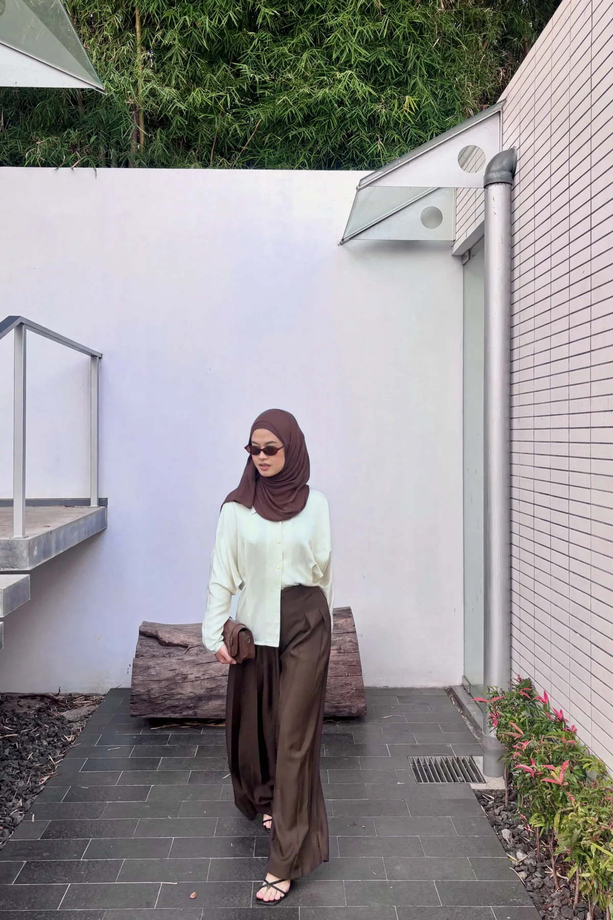 Woman in cream silk blouse and brown pleated trousers with camel hijab leaning on a brick wall