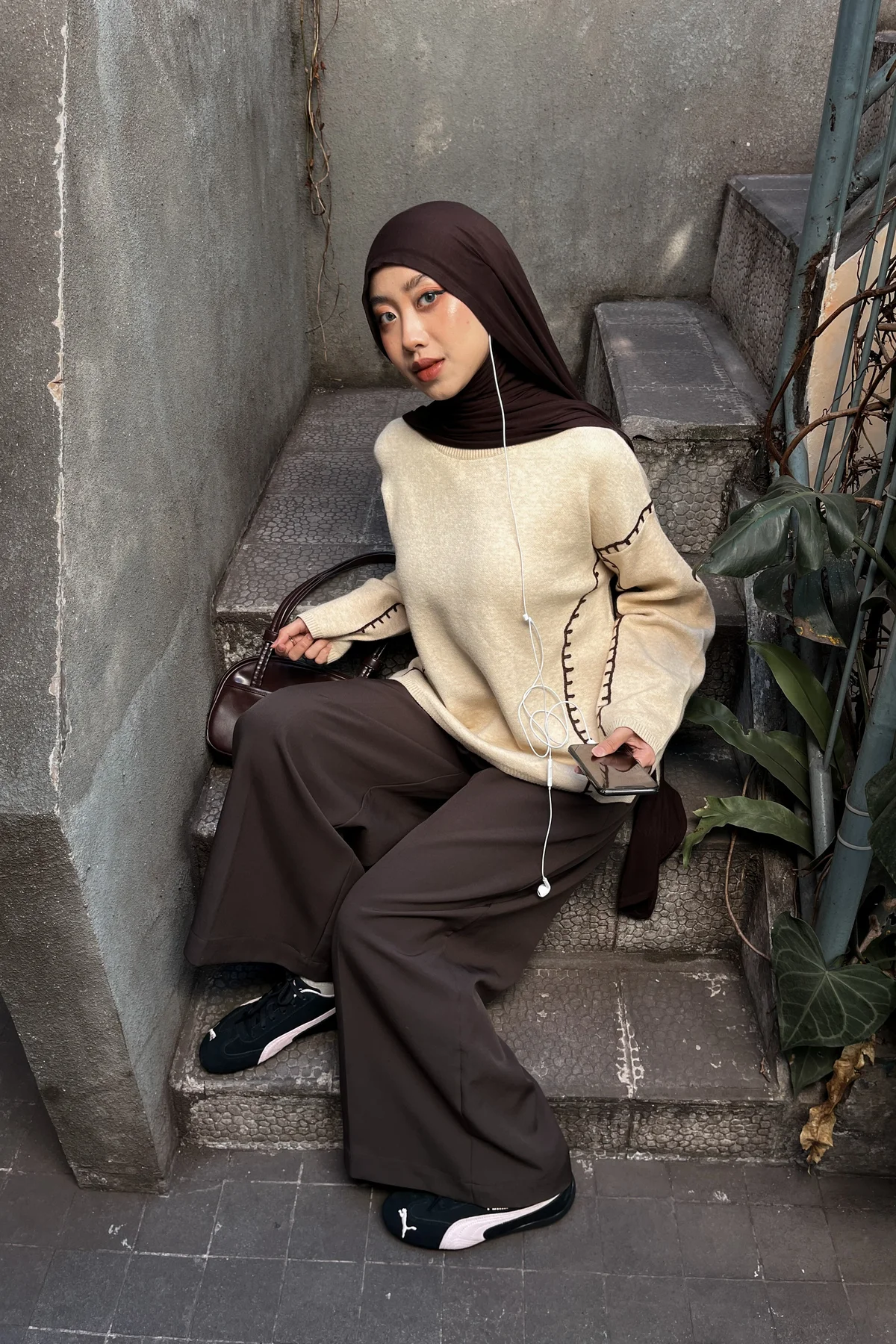 Hijabi woman in cream knit and charcoal track pants walking a park path, casual Christmas outfit