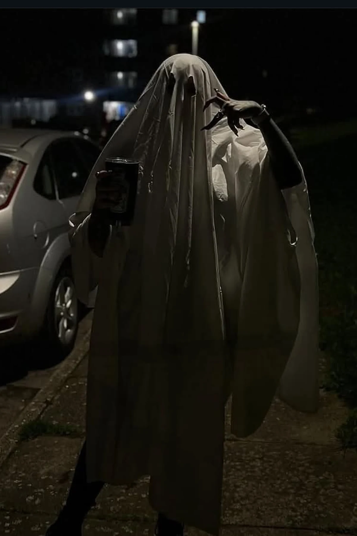 Person in a white sheet ghost costume with dark eye holes at night, a DIY sheet ghost Halloween costume