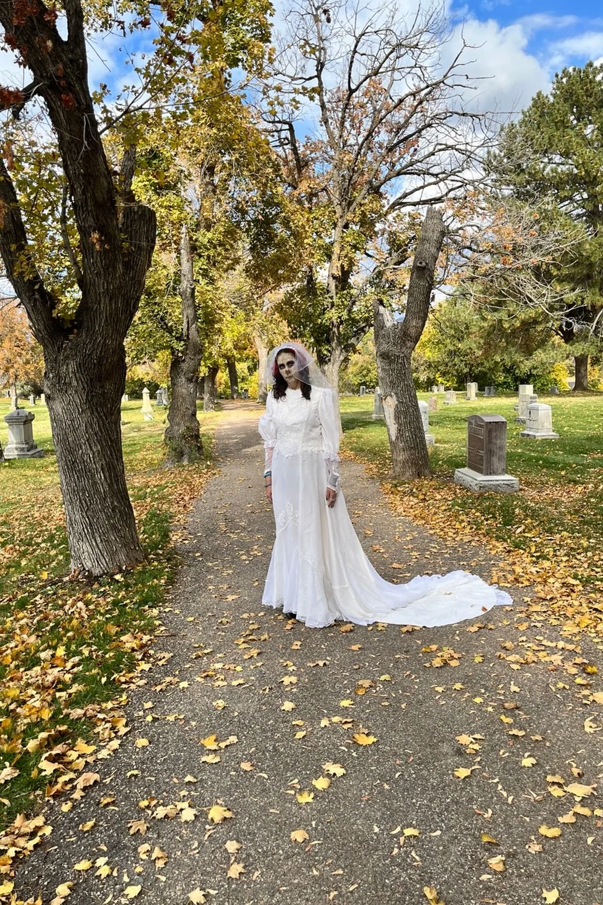 Woman in her 50s in a tattered wedding gown and torn veil holding dead black roses, a zombie bride solo horror Halloween costume