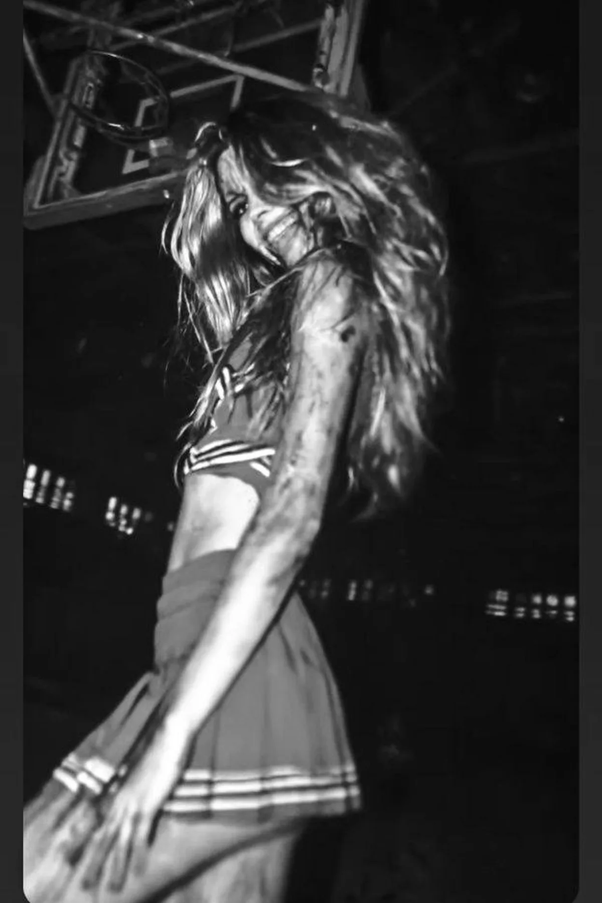 Woman in her 50s in a tattered cheerleader uniform with ragged pom-poms, a zombie cheerleader solo horror Halloween costume