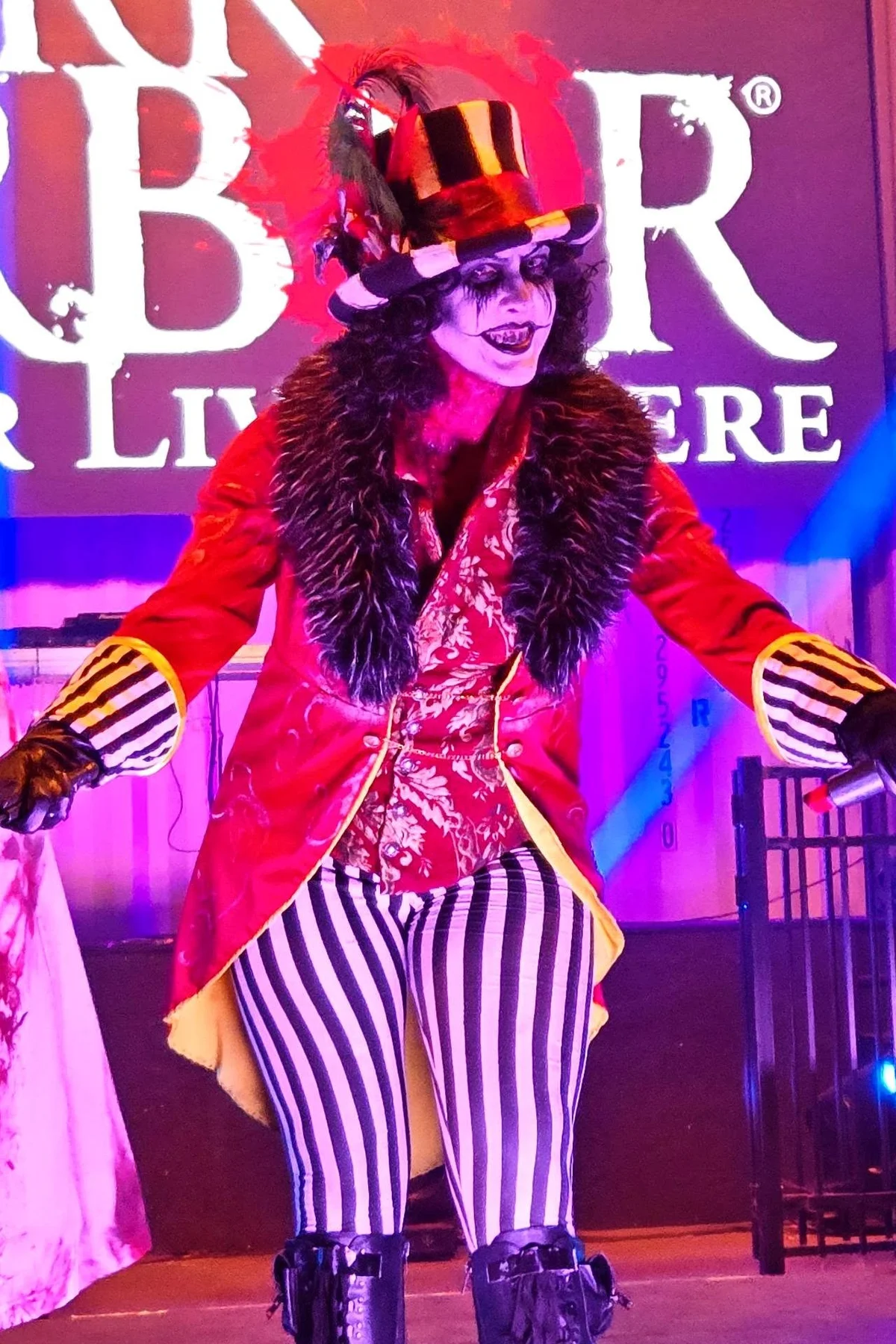 Woman in her 50s in a red tailcoat and striped top hat with skull-face makeup holding a whip, a haunted ringmaster solo horror Halloween costume