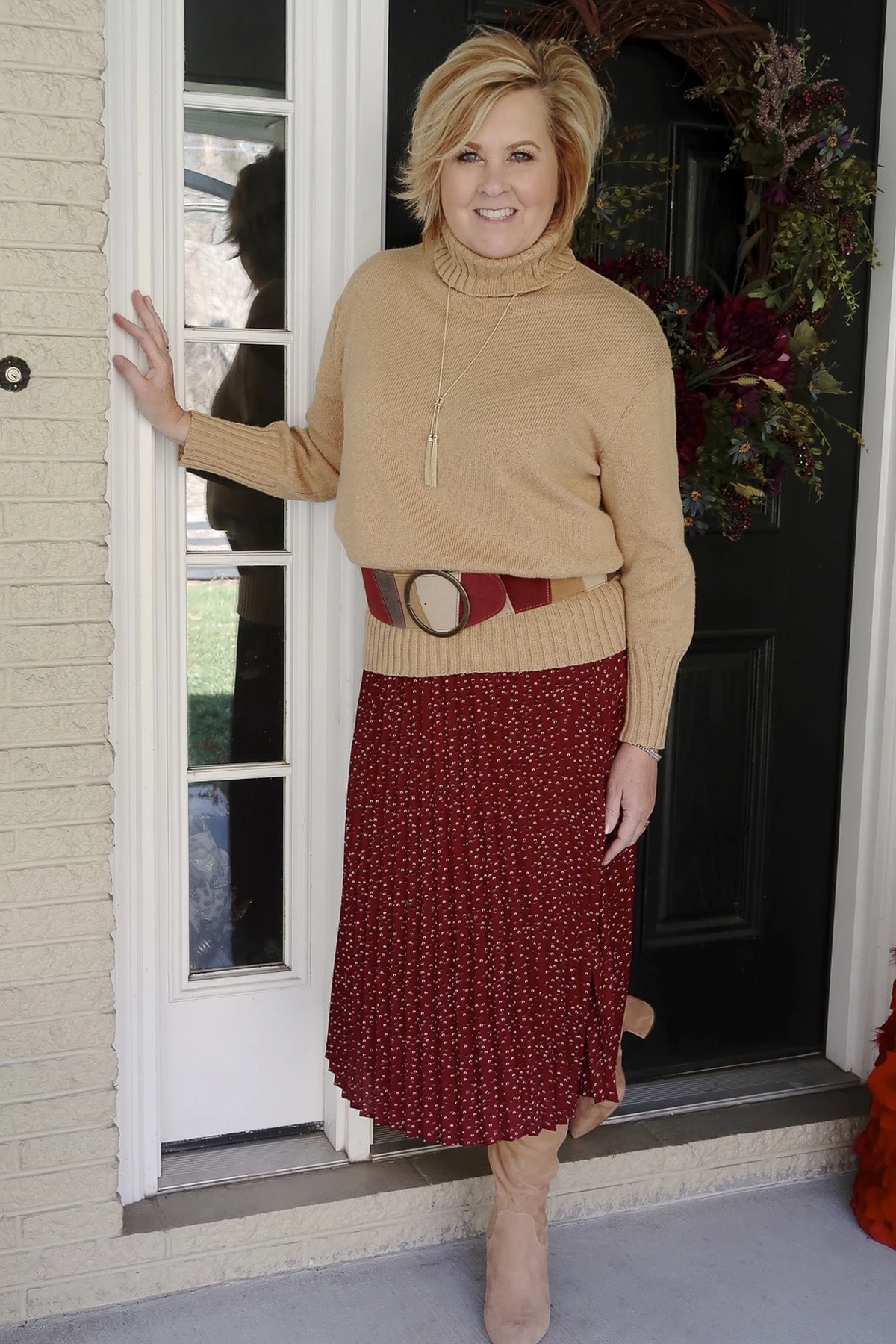 Woman in camel turtleneck and burgundy pleated skirt on a porch with a wreath