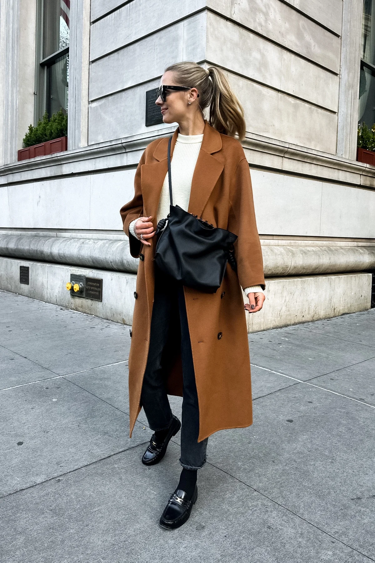 Woman in camel coat, ivory knit sweater and black jeans on a city street