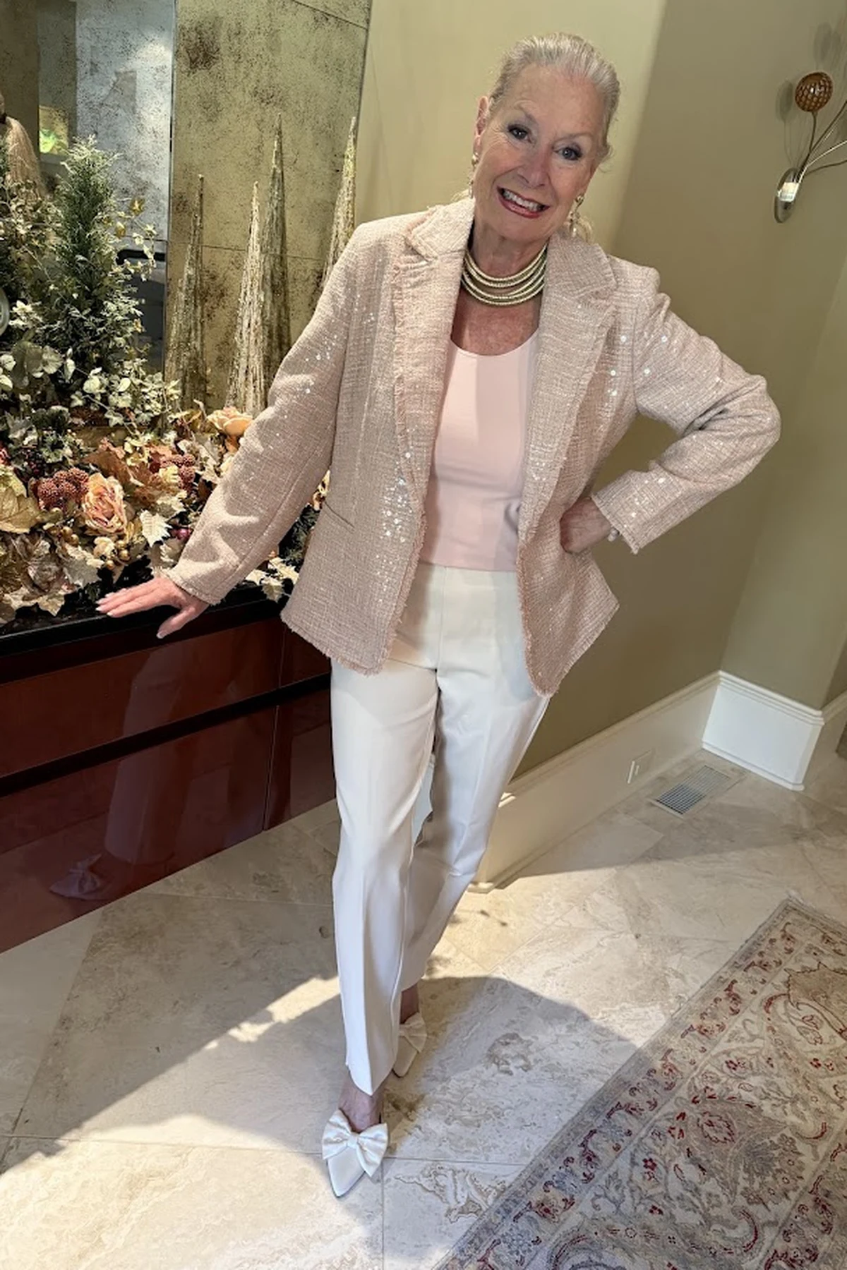 Woman in champagne sequin blazer and white trousers beside Christmas greenery