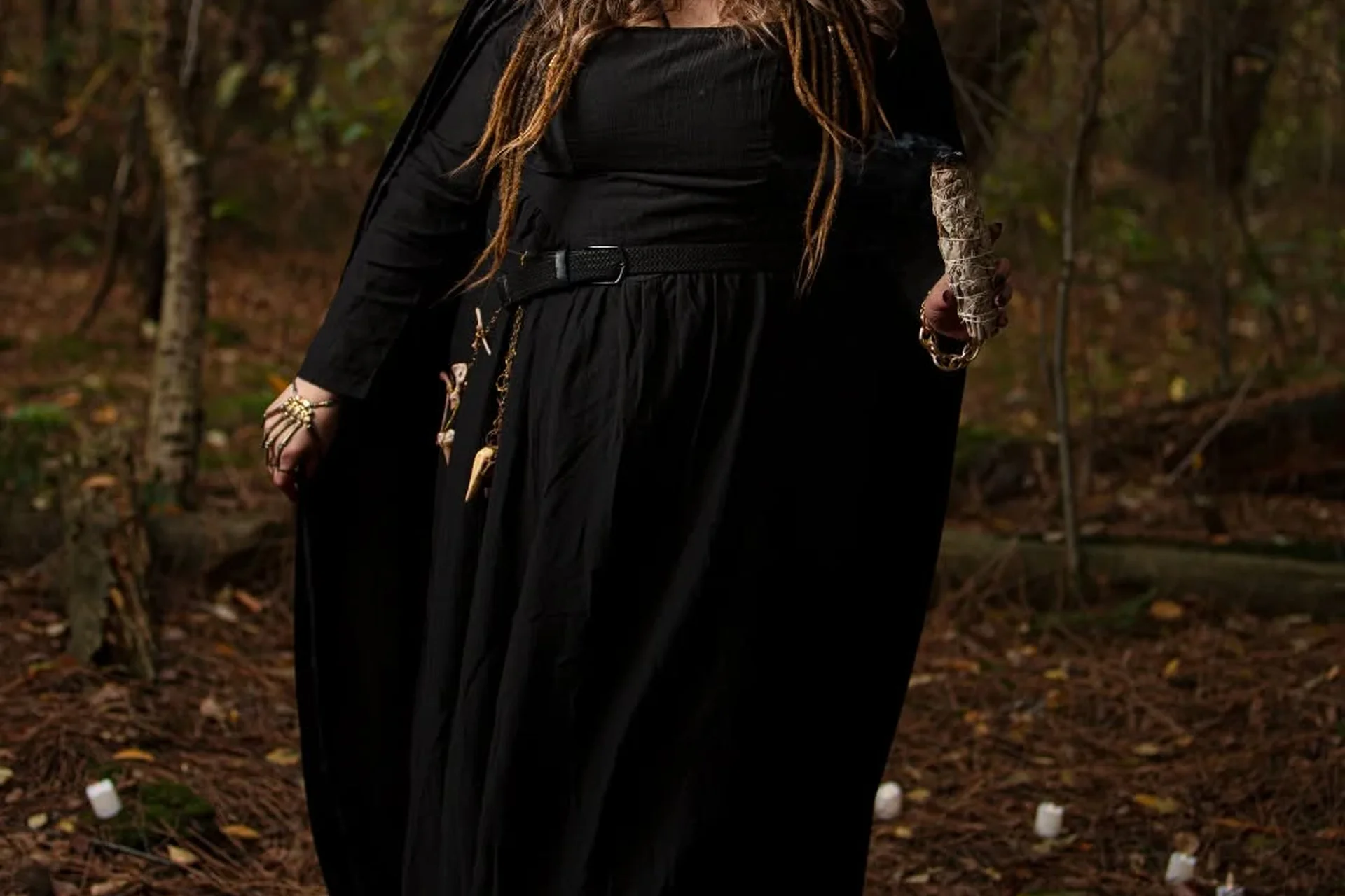 Plus size woman in a flowing black hooded cloak holding a smoldering sage bundle in a candlelit forest, a gothic Halloween hero look