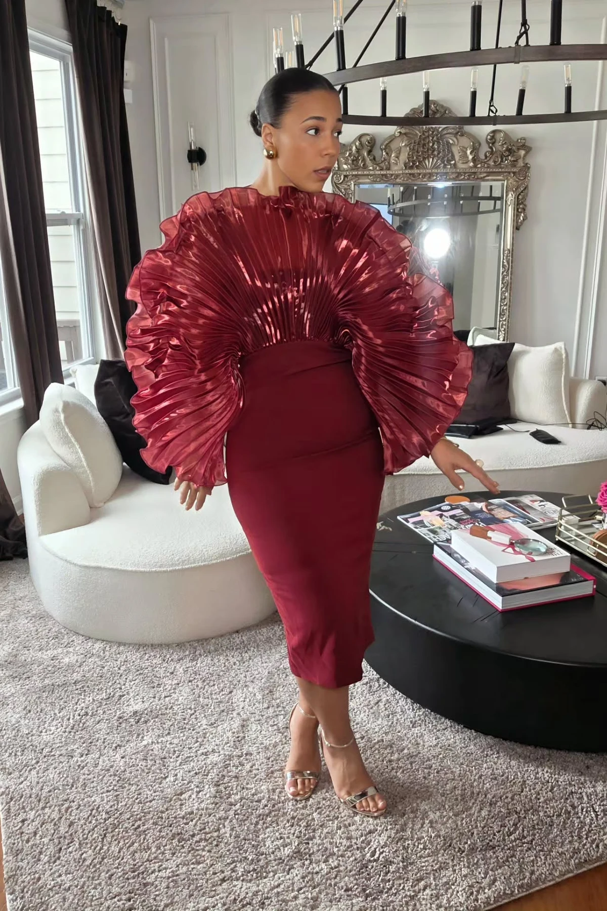 Woman in a burgundy midi dress with pleated fan statement sleeves in a chandelier-lit living room