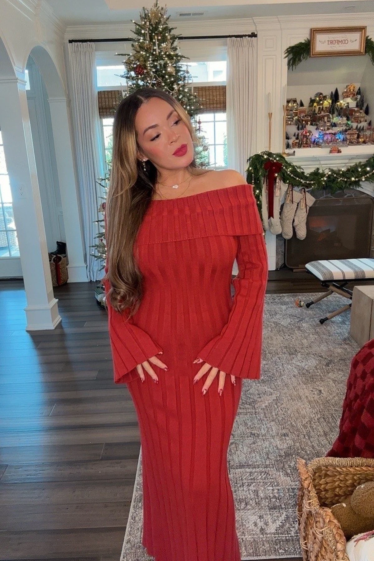 Woman in a red off-the-shoulder knit maxi dress with bell sleeves by a Christmas tree and fireplace