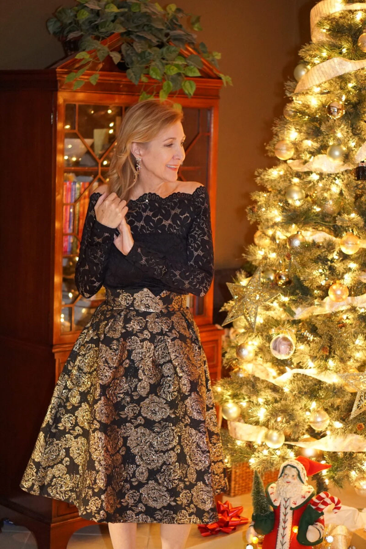 Woman in a black lace long-sleeve top and black-and-gold brocade skirt beside a lit Christmas tree