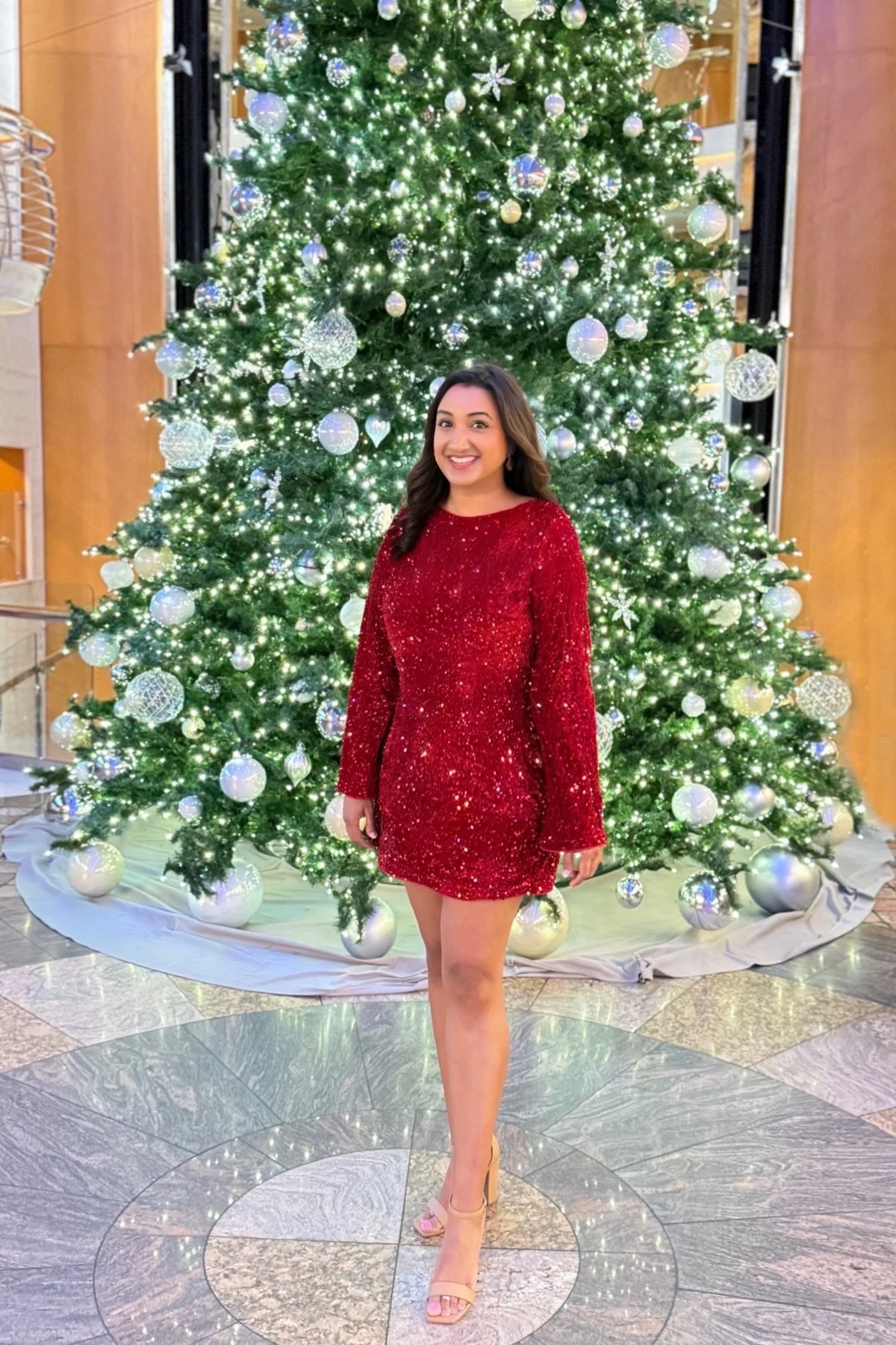 Woman in a red sequin long-sleeve mini dress with nude heels in front of a giant decorated Christmas tree