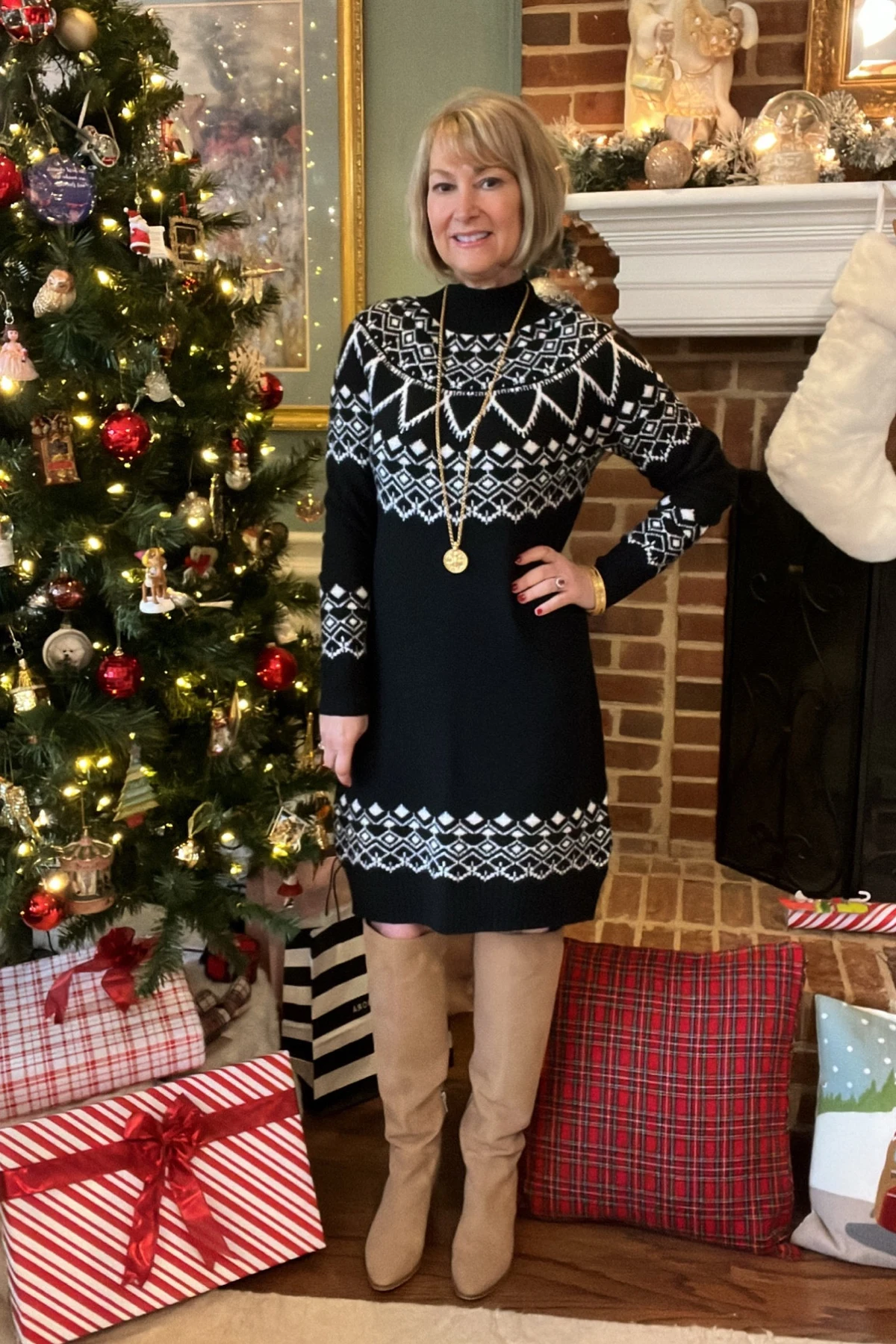 Woman over 50 in a black Fair Isle sweater dress with long sleeves by a Christmas tree and fireplace