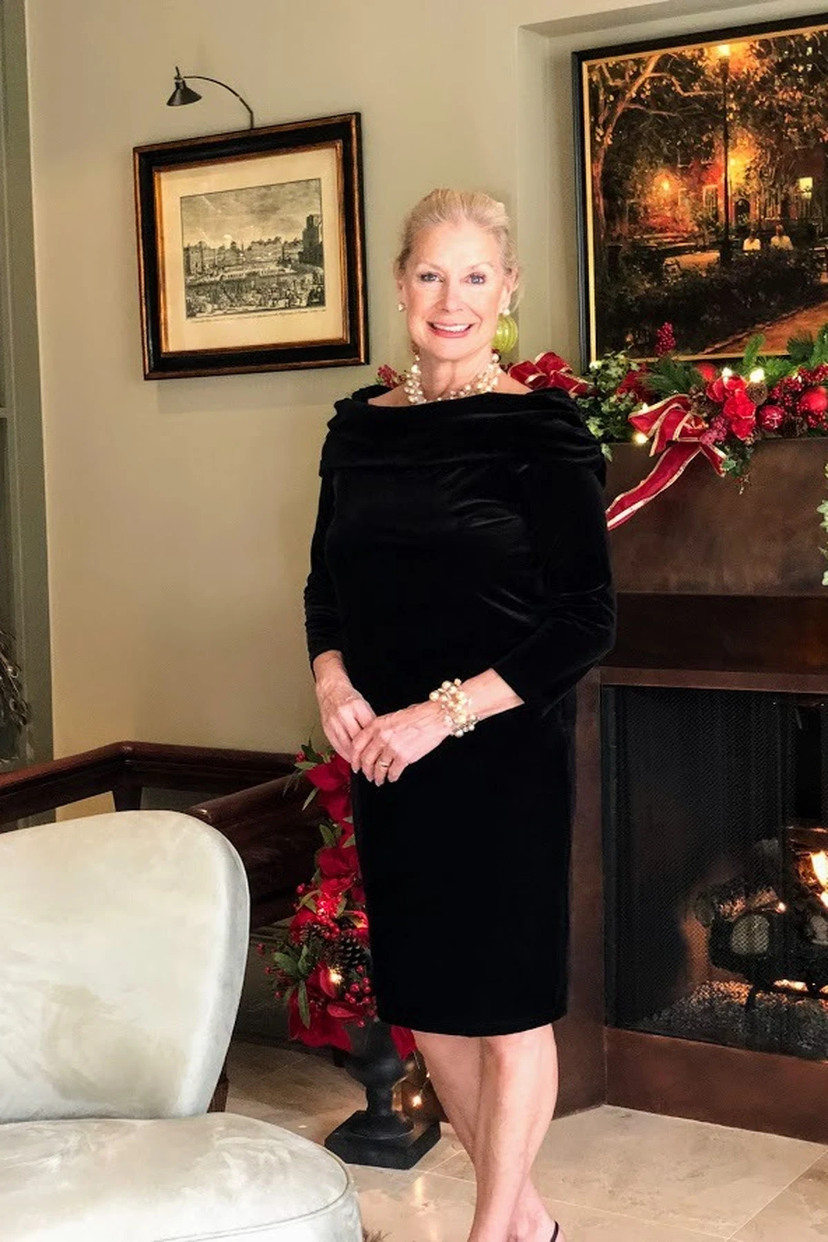 Woman over 50 in a black velvet off-the-shoulder sheath dress with three-quarter sleeves by a garland and fireplace