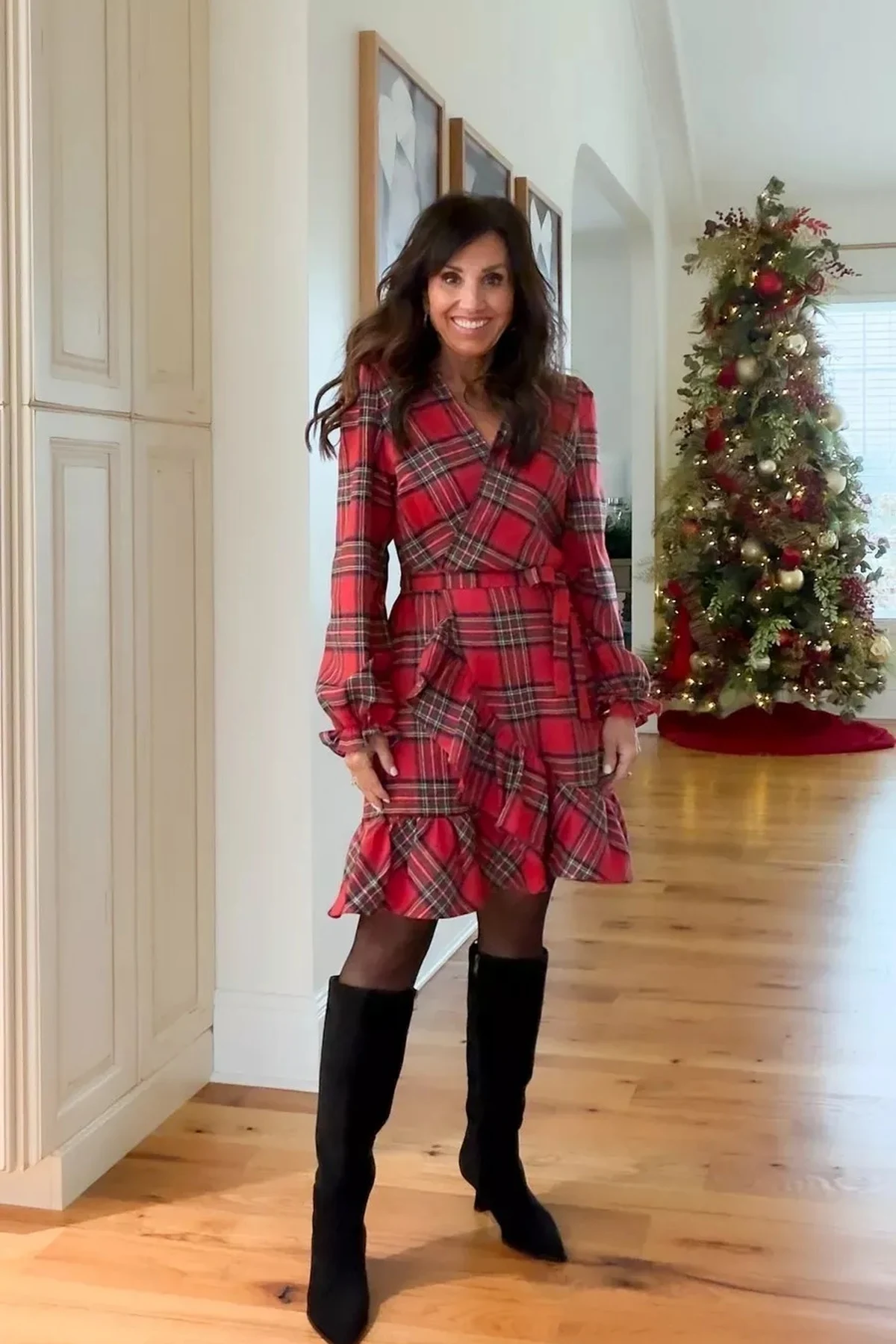 Woman in a red tartan plaid wrap dress with long blouson sleeves and tall boots by a Christmas tree