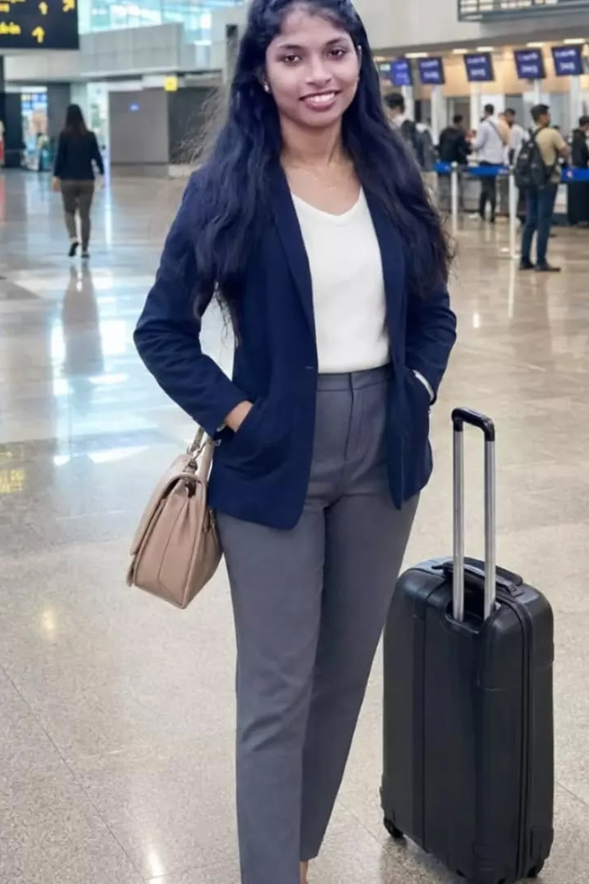 Woman in navy knit blazer, cream turtleneck and black trousers pulling a carry-on at an airport